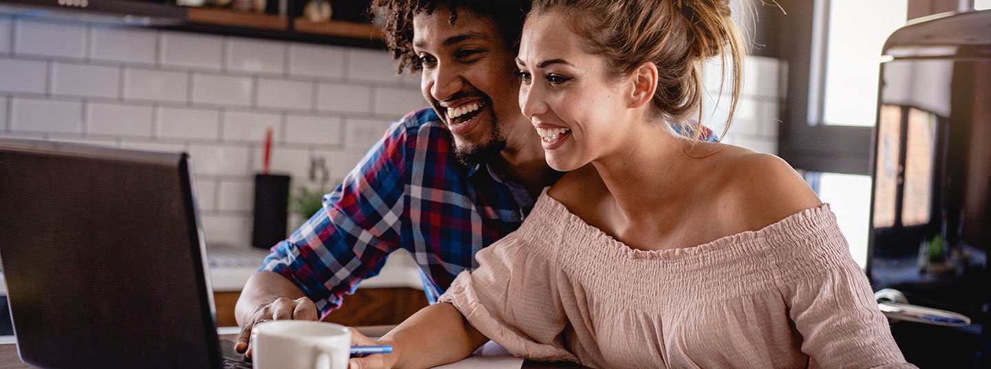 A smiling man and woman sit close together at a table, sharing a joyful moment while looking at a laptop.